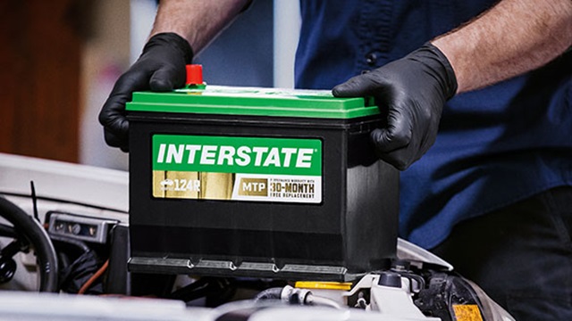 Main photo for Interstate Batteries page on ByrneAuto.com depicting the gloved hands of an auto technician placing a new Interstate battery under the hood of a car.